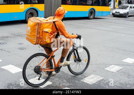 Fast-Food-Lieferung männlichen Kurier trägt die Take-away-Firma orange Outfit mit Fahrrad auf belebten Straße in Sofia Bulgarien Stockfoto