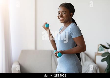 Heimsport. Fröhliche afrikanisch-amerikanische Lady beim Training mit Hanteln im Wohnzimmer Stockfoto