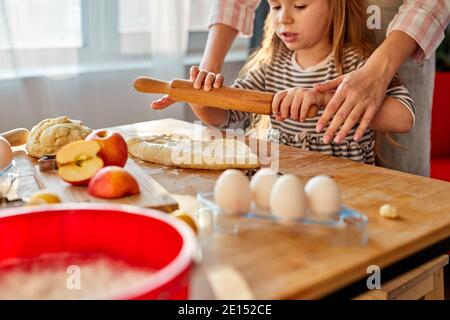 Die junge Mutter lehrt ihre fleißige Tochter zu kochen, steht hinter dem Tisch und rollt den Teig aus Stockfoto