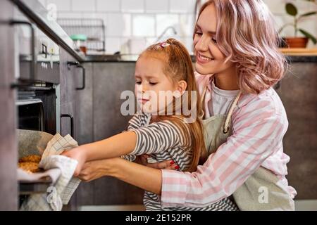 Mutter und nettes Kind Tochter backen Cookies in der Küche, lächelnd kaukasischen Mutter Unterricht Kind nehmen Tablett mit Cookies aus Ofen Herd kochen zu Hause, famii Stockfoto