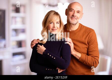 Ein glückliches Paar mittleren Alters, das sich umarmte, während es zu Hause im Wohnzimmer zusammenstand. Stockfoto