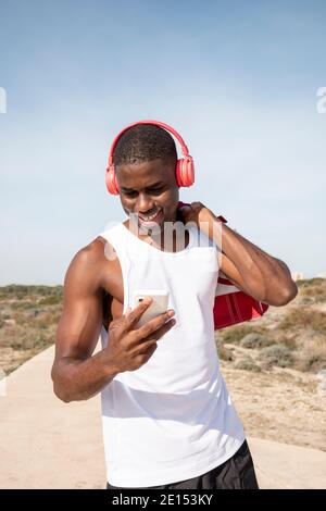 Smiley junger schwarzer Sportler mit Kopfhörern, der mit seinem Handy Musik hört. Konzept der Technologie. Stockfoto