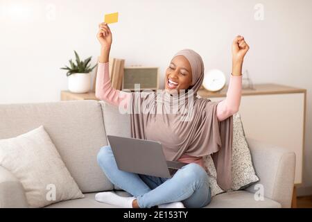 Schwarze Frau in Hijab glücklich über riesigen Rabatt im Online-Shop, Kauf von Waren zum Verkauf, mit Laptop und Kreditkarte zu Hause Stockfoto