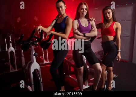 Athlet Damen stehen zusammen in der Nähe von stationären Bikes im Fitnessstudio, posiert vor der Kamera. Isoliert in rot neon beleuchteten Raum im Hintergrund Stockfoto