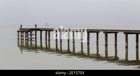 Holzsteg am Ammersee an einem nebligen Wintertag. Im Hintergrund ein Segelboot. Stockfoto