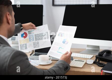 Beschnittene Ansicht des Traders, der Papiere mit Charts in der Nähe von Computermonitoren und Kaffeetasse betrachtet, verschwommener Vordergrund Stockfoto