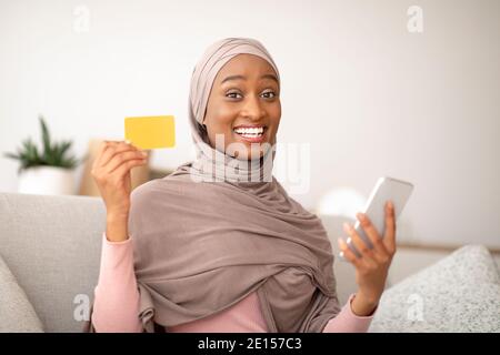 Glückliche schwarze Frau in Hijab hält Kreditkarte und Handy, Kauf von Waren online über Anwendung zu Hause Stockfoto