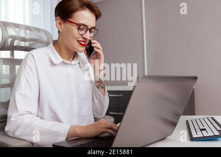 Attraktive junge Freiberuflerin, die am Laptop arbeitet und ihr Smartphone benutzt. Weibliche IT-Industriearbeiterin, die Computer verwendet Stockfoto