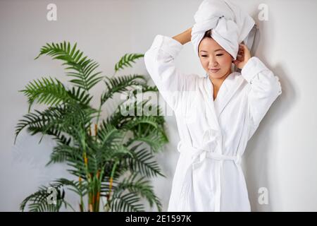 Mixed Race Frau nach dem Bad, trägt Handtuch und Bademantel, steht in der Nähe grüne Pflanze in hellen Raum Stockfoto