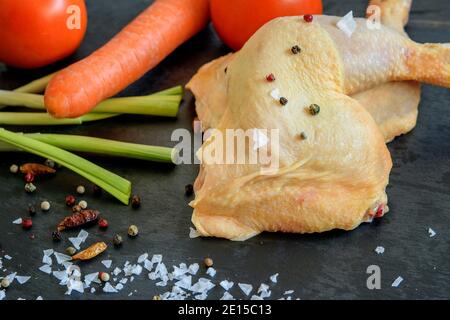 Rohe, ungekochte Hähnchenschenkel mit Gemüse und Gewürzen auf dem schwarzen Kücheboard, das zum Kochen vorbereitet ist. Gesunde Ernährung Konzept. Stockfoto