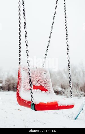 Verschneite Babyschaukel im Winter - leerer Spielplatz - Roter Plastikschaukelstuhl in der Kälte Stockfoto