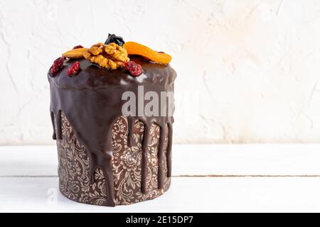 Osterkuchen oder Kulitch mit glasiert mit Schokolade und verziert mit Nüssen, getrockneten Beeren und Früchten auf weißem Holztisch. Stockfoto