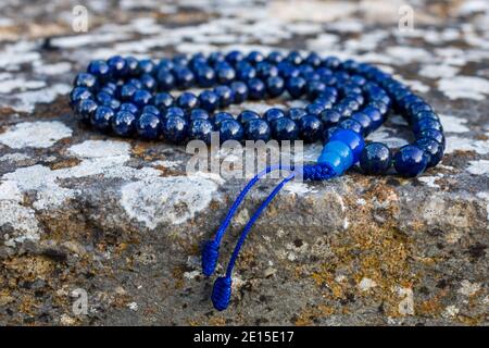 Blaue mala Perlen (Lapislazuli) an Steinwand, Nahaufnahme und selektiver Fokus. Achtsamkeit und Meditationszubehör. Stockfoto