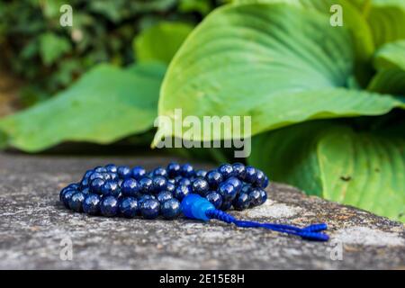 Blaue mala Perlen auf Steinwand mit grünen Blättern im Hintergrund, selektiver Fokus. Yoga und Meditation Zubehör. Stockfoto