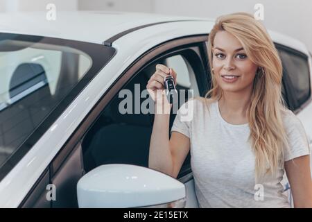 Wunderschöne langhaarige blonde lächelnd, halten Autoschlüssel bei der Händler. Glückliche Frau posiert mit ihrem neuen Auto. Weibliche Fahrerin, die sich auf ihr vehi stützt Stockfoto