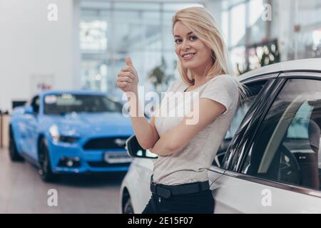 Aufgeregt junge schöne weibliche Fahrerin posiert in der Nähe ihres neu gekauften Autos im Autohaus, zeigt Daumen nach oben, Copy Space. Wunderschöne Frau beim Kauf Stockfoto