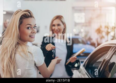 Wunderschöne junge Frau lächelt auf die Kamera über ihre Schulter, während sie die Autoschlüssel für ihr neues Auto im Autohaus erhält. Verkäuferin, die den Schlüssel überreicht Stockfoto