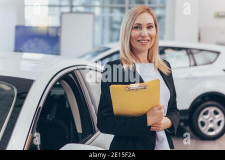 Professioneller Autohändler posiert stolz im Auto Showroom, lächelt an der Kamera, hält Zwischenablage. Glückliche Verkäuferin im Autohaus, Stockfoto