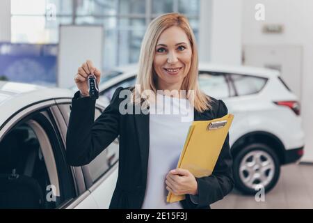 Reife Autohändlerin lächelt fröhlich zur Kamera, hält die Autoschlüssel, steht vor einem neuen Auto im Verkauf. Verkäuferin, die im Auto arbeitet Stockfoto