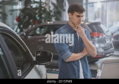 Junge attraktive Mann Blick auf neue Auto weit Verkauf nachdenklich, reiben sein Kinn. Schöner männlicher Kunde, der neues Automobil im Händlersalon wählt. T Stockfoto