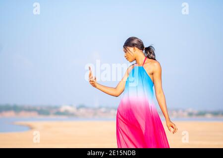 Hübsche junge afrikanische Dame, die ein farbenfrohes Kleid am Strand trägt, ihr Handy benutzt und ein Selfie-Foto von ihrem Urlaub macht, um es in den sozialen Medien zu posten Stockfoto