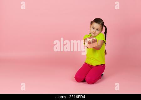 Mock up Mädchen mit Zöpfen in einem hellgrünen T-Shirt und roten Hosen reicht uns eine Geschenkbox auf dem Boden sitzen , isoliert auf einem rosa Hintergrund Stockfoto