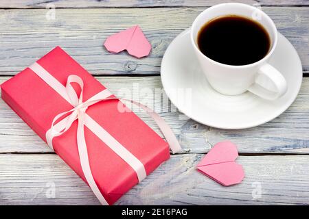 Ein rosa Geschenk mit einem weißen Band gebunden, eine Tasse Kaffee und Herzen auf einem hellen Holztisch. Stockfoto