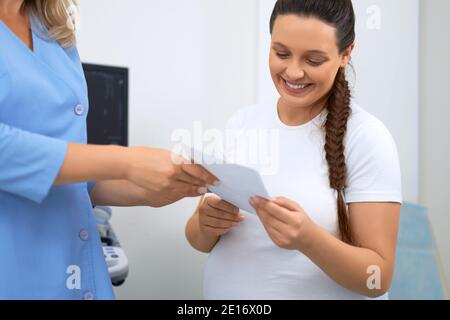 Lächelnde junge Frau in Erwartung Blick auf Foto nach Ultraschall-Screening. Ärztin, die professionelle Beratung für zukünftige Mutter in der Klinik. Stockfoto