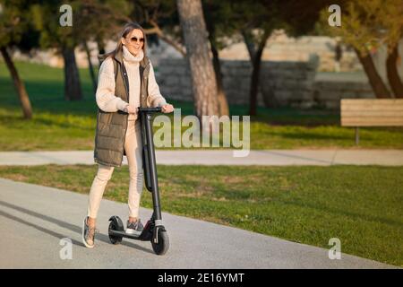 Junge stilvolle Frau Reiten Elektroroller Kick im Stadtpark. Elektrischer Öko-Transport Stockfoto