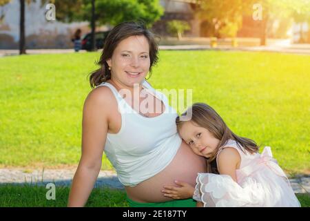 Nettes kleines Mädchen umarmt und küsst und hört ihre Mütter schwangeren Bauch sitzen im Freien auf einer grünen Wiese in einem Stadtpark auf einer sonnigen da Stockfoto