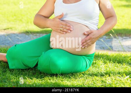 Junge schwangere Frau, die ihren Magen vor dem Training mit Sonnenschutzcreme schützt, während sie im Park auf grüner Wiese sitzt. Erwartete Frau, die CR anwendet Stockfoto