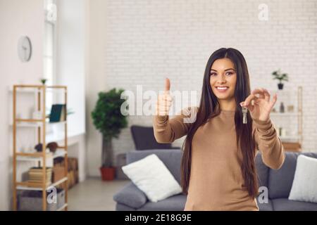 Glückliche junge Frau mit den Schlüsseln zu ihrer neuen Wohnung, lächelnd und Daumen nach oben Stockfoto