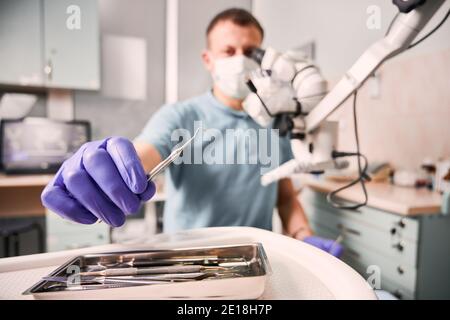 Nahaufnahme der männlichen Zahnarzt Hand in sterilen Handschuh hält Metall Dental Explorer. Stomatologe in der medizinischen Maske unter zahnärztliche Instrument, während in der Nähe von diagnostischen Mikroskop sitzen. Konzept der Zahnmedizin. Stockfoto