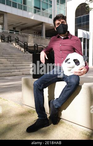 Der junge Geschäftsmann trägt eine Gesichtsmaske mit einer Panda-Kopfmaske in den Händen und einen Koffer, sitzt auf einer Bank. Bizarrer Geschäftsmann. Stockfoto