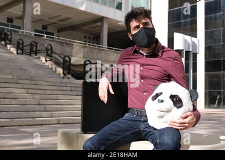 Der junge Geschäftsmann trägt eine Gesichtsmaske mit einer Panda-Kopfmaske in den Händen und einen Koffer, sitzt auf einer Bank. Bizarrer Geschäftsmann. Stockfoto