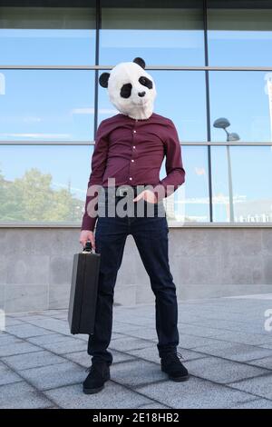 Junger Geschäftsmann, der eine Panda-Kopfmaske trägt und einen Koffer vor den Firmengebäuden trägt. Bizarrer Geschäftsmann. Stockfoto