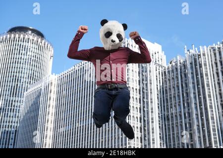 Der junge Geschäftsmann trägt eine Panda-Kopfmaske und springt vor die Firmengebäude. Bizarrer Geschäftsmann. Stockfoto