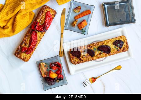 Hausgemachter gesalzener Kuchen mit Gemüse und gerösteten Paprika darauf. Rechteckige Formen auf weißem Marmorboden. Untertassen aus Glas, goldenes Besteck und ein Handtuch. Hochwertige Fotos, Draufsicht Stockfoto