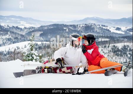 Charmante junge Frau, die den Mann anschaut und lächelt, während sie den Arm um seine Schulter wickelt. Fröhlicher Freund und Freundin sitzen auf schneebedeckten Hügel in den Bergen. Konzept des Skifahrens und Beziehungen. Stockfoto