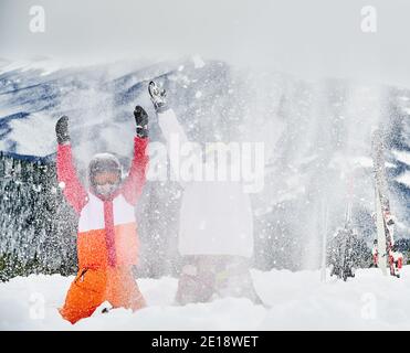 Zwei Skifahrer in Skianzügen und Helmen werfen frischen Pulverschnee hoch in die Luft, Spaß im Skigebiet mit Bergen im Hintergrund. Konzept der Wintersport-Aktivitäten, Spaß und Beziehungen. Stockfoto