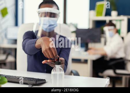 Nahaufnahme einer afrikanischen Frau, die während einer globalen Pandemie mit Coronavirus Handdesinfektionsmittel verwendet. Geschäftsfrau in neuen normalen Arbeitsplatz desinfizieren, während Kollegen arbeiten im Hintergrund. Stockfoto