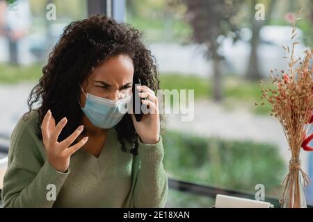 Wütend afroamerikanische Frau in medizinische Maske im Gespräch auf dem Smartphone Und gestikulieren im Café Stockfoto