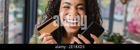 Fröhliche afroamerikanische Frau lachend, während sie Smartphone und Kreditkarte, Banner hält Stockfoto