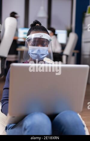 Afrikanische Geschäftsfrau arbeiten auf Laptop tragen Gesichtsmaske gegen covid19. Multiethnisches Geschäftsteam, das während der globalen Pandemie mit Coronavirus soziale Distanz respektiert. Stockfoto