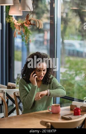 Verärgert afroamerikanische Frau sprechen auf dem Smartphone beim Überprüfen Uhr Am Handgelenk und im Café warten Stockfoto