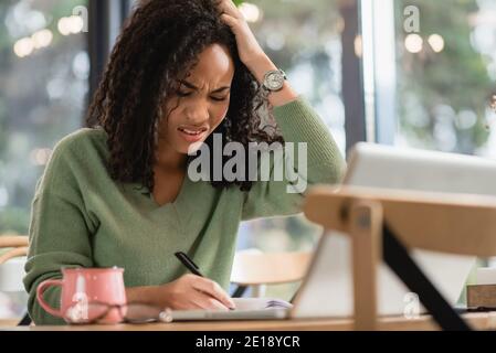 Erschöpft afroamerikanische Frau schriftlich in Notebook während des Studiums online Im Café Stockfoto