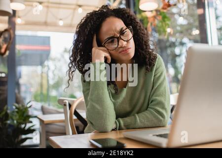 Nachdenkliche afroamerikanische Frau in der Brille Blick auf Laptop in der Nähe Smartphone auf dem Tisch Stockfoto