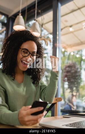 Aufgeregt afroamerikanische Frau in der Brille Blick auf Smartphone auf Verschwommener Vordergrund Stockfoto