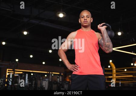 Low-Angle-Aufnahme eines muskulösen afrikanischen männlichen Athleten, der selbstbewusst auf die Kamera schaut und die Kugelhantel hält. Fokussierter Crossfit-Sportler, der stolz posiert Stockfoto