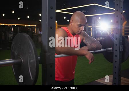 Muskulöser, afrikanischer Mann, der sich nach dem Training mit schwerer Langhantel ausruhte. Attraktiver junger Bodybuilder, der sich auf eine Langhantel stützt und nach dem Aufstehen müde aussieht Stockfoto